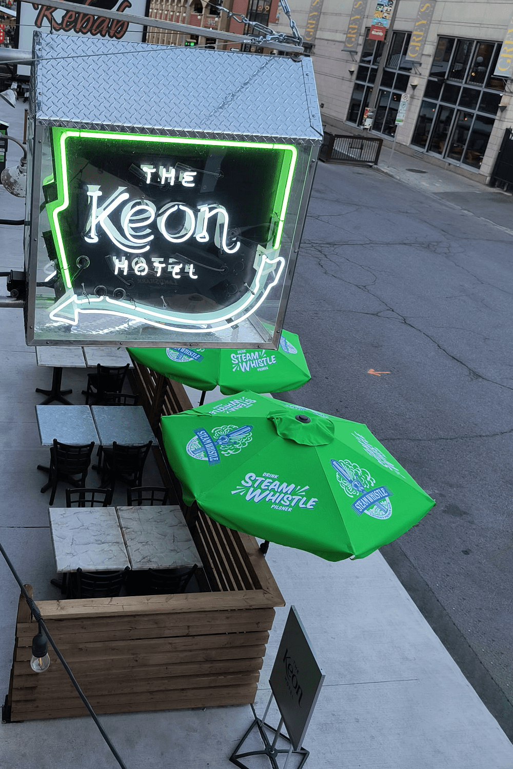 Neon sign for "The Keon Hotel" above a patio with bright green umbrellas labeled "Steam Whistle." Empty tables and chairs, street in the background.