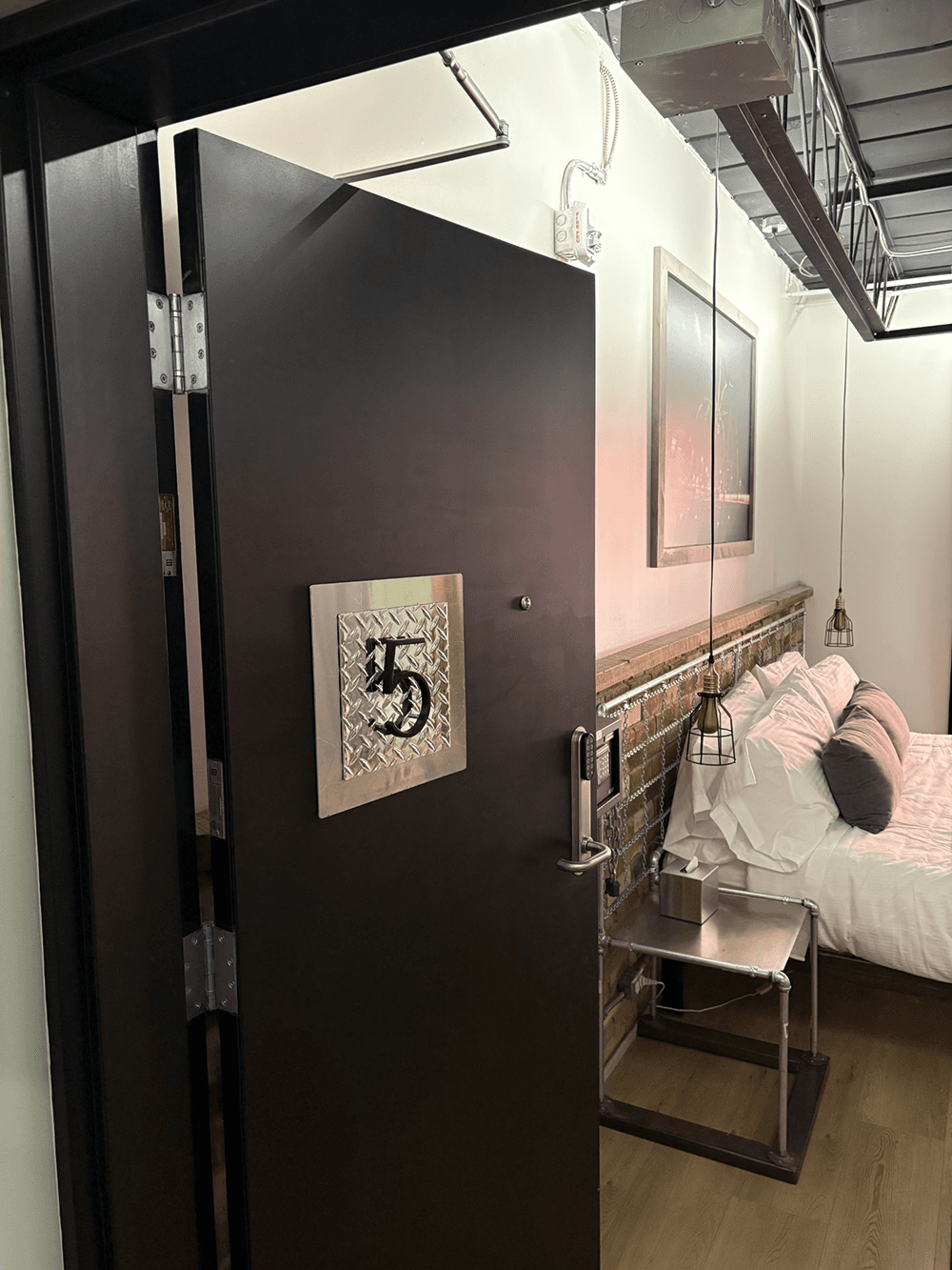 A black door with a metallic plaque displaying the number 45 opens into a modern bedroom with a white bed and industrial decor.