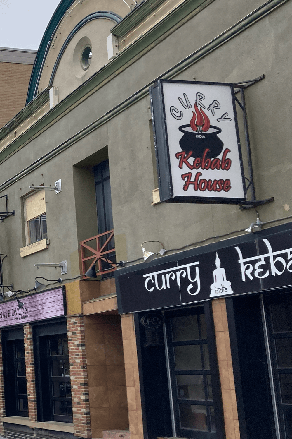 Street view of a building with a 'Curry Kebab House' sign featuring a steaming pot. Nearby shop has a closed, rustic facade. Overcast atmosphere.
